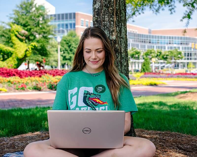 Female student wearing a green shirt with Blaze on it, sitting under tree with a laptop.